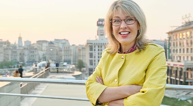 Oral-Surgery-Bucket Middle-aged woman in glasses and yellow jacket smiling on a city balcony