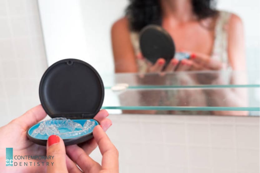 Clear Invisalign aligners in a case being held in front of a bathroom mirror