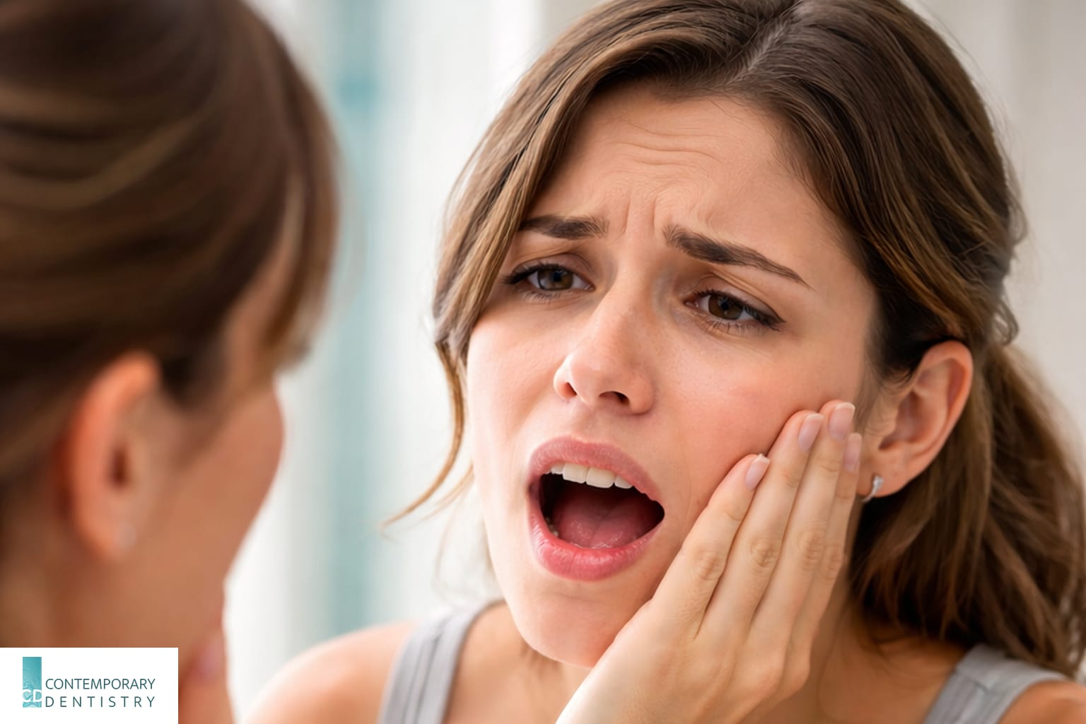 woman-toothache-looking-in-mirror Woman touching her cheek while looking in a bathroom mirror with a painful expression, illustrating toothache or dental pain.